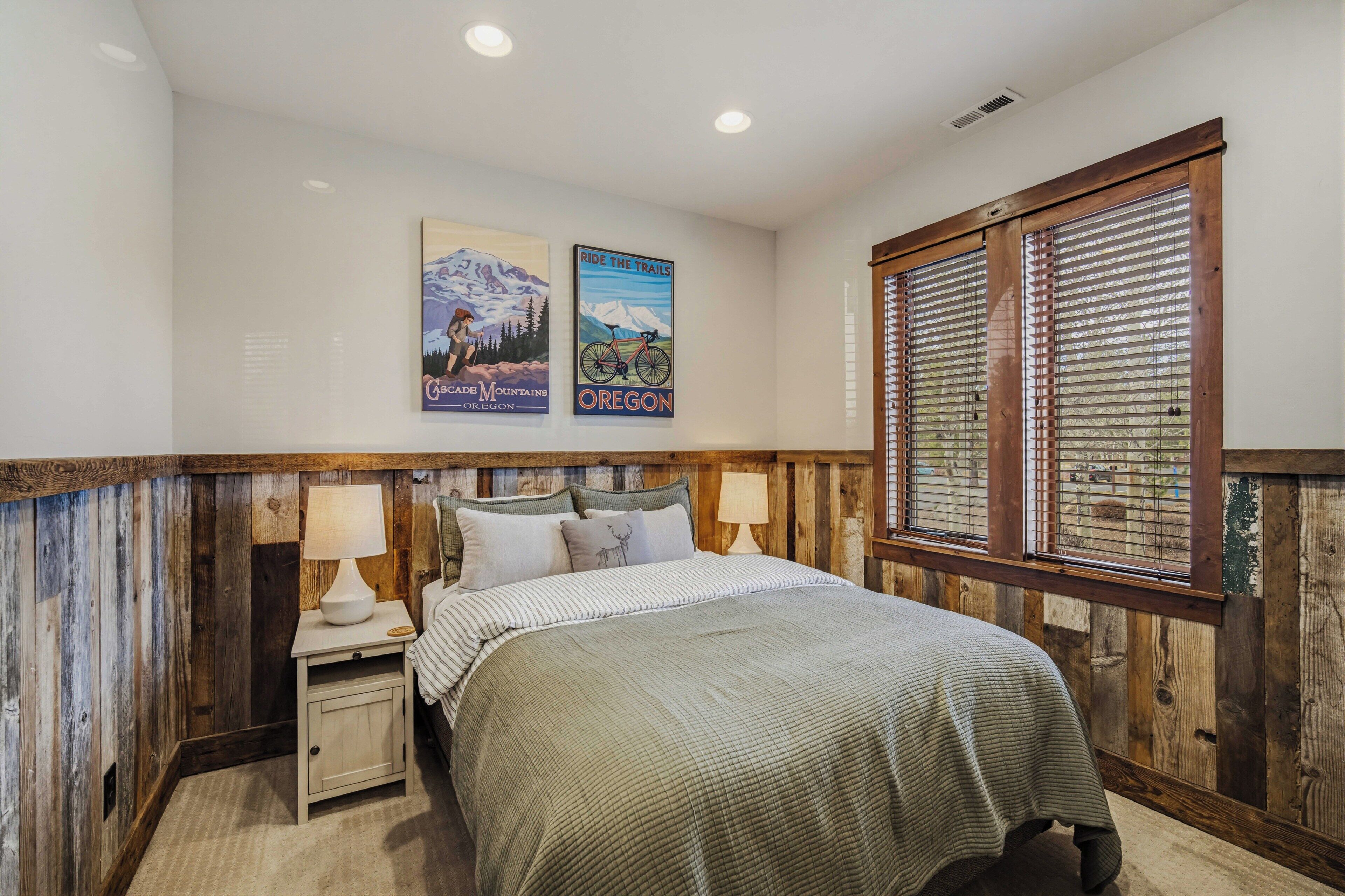 A bedroom with a bed, two bedside tables with lamps, wooden paneling, a window with blinds, and two wall posters.