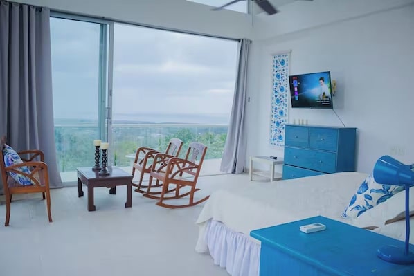 Una habitación de hotel moderna con un ventanal grande, una cama, un escritorio, un televisor y un aparador azul.