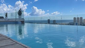 Cielo azzurro e limpido con nuvole, piscina con bordo in legno ed edifici moderni sullo sfondo.