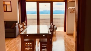 A spacious room with a wooden dining table, a sofa, and a large window offering a view of mountains.