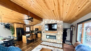 A spacious living room with a stone fireplace, wooden ceiling, and a large TV mounted on the wall.