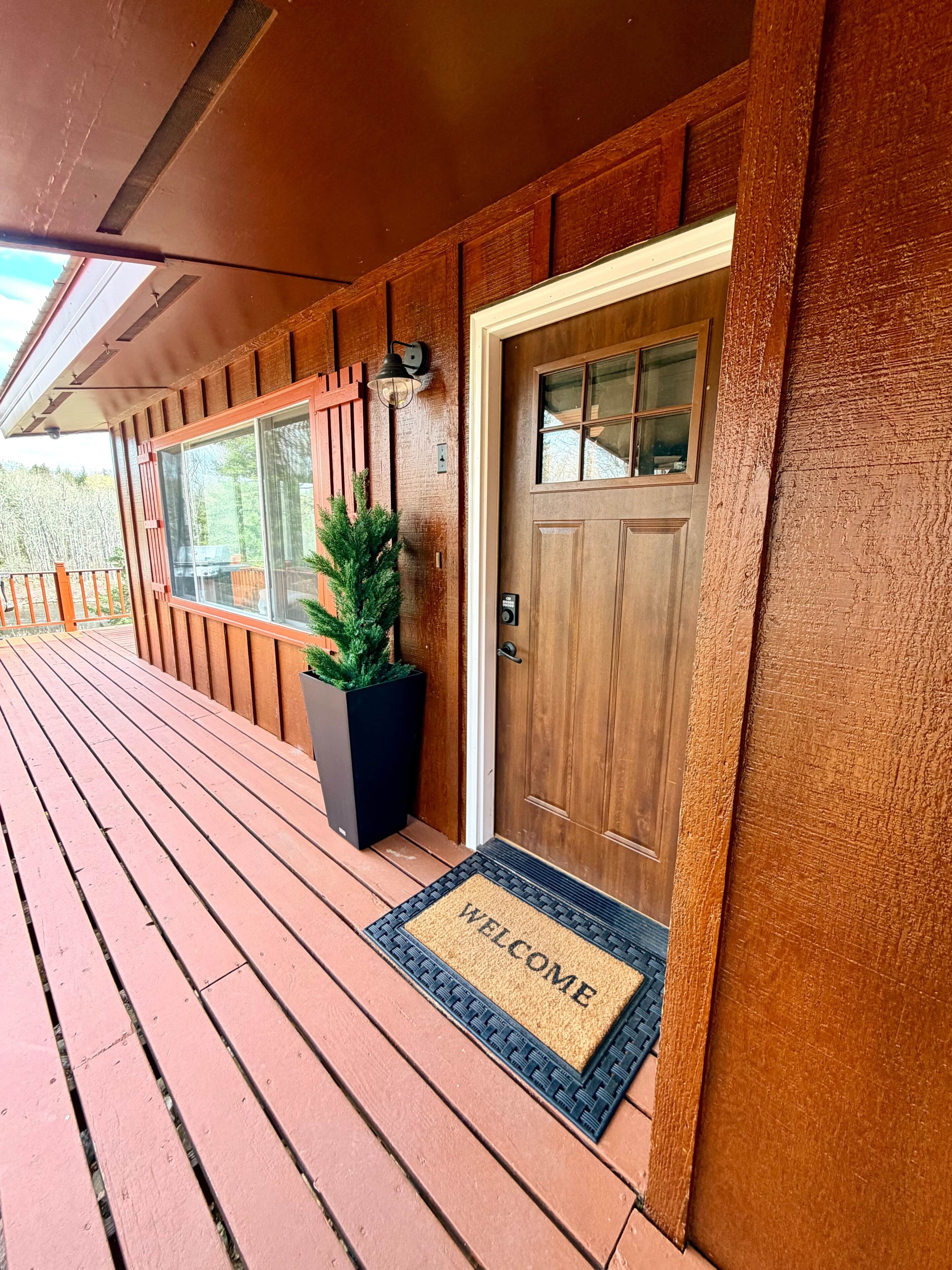 Une terrasse en bois avec un paillasson, une plante en pot et une porte dotée d’une fenêtre.