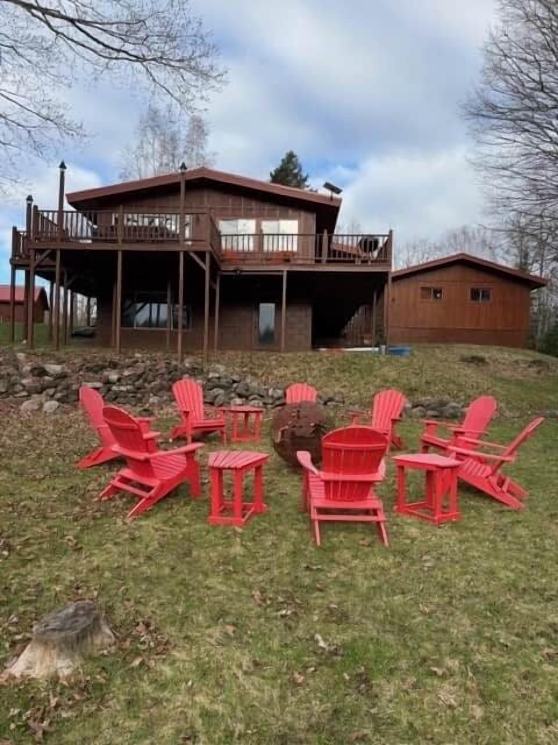Eine Holzhütte mit einem Balkon, ein separates Gebäude und rote Adirondack-Stühle auf einer Rasenfläche.