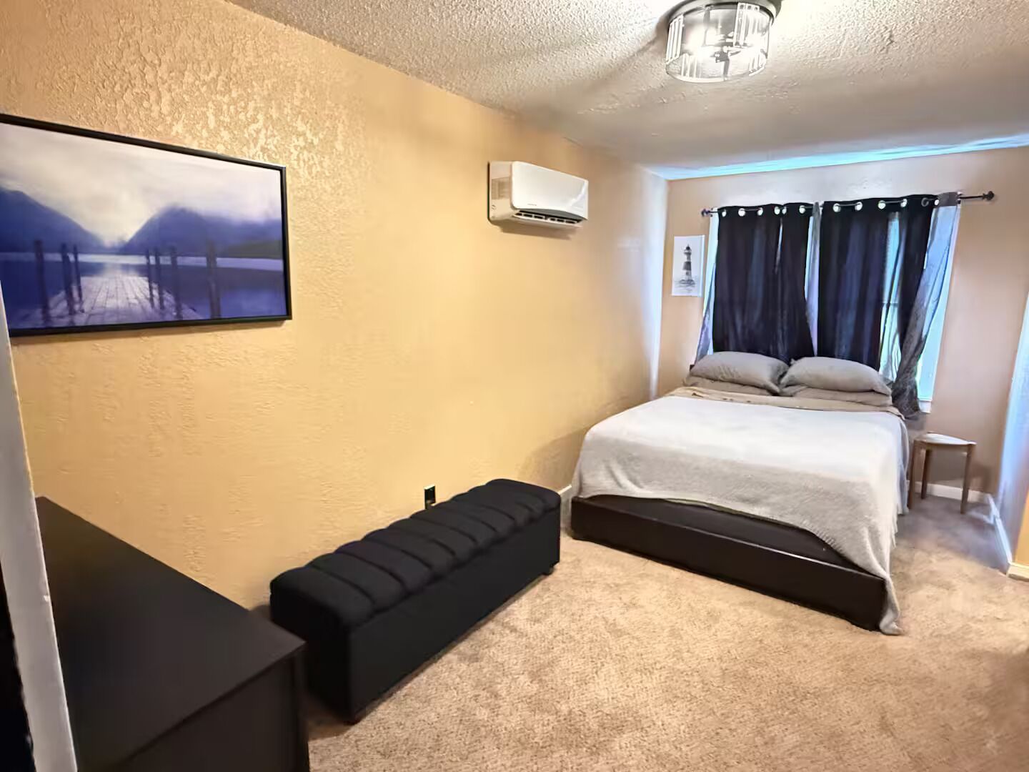 A bedroom with a bed, a bench, an air conditioner, and a painting on the wall.