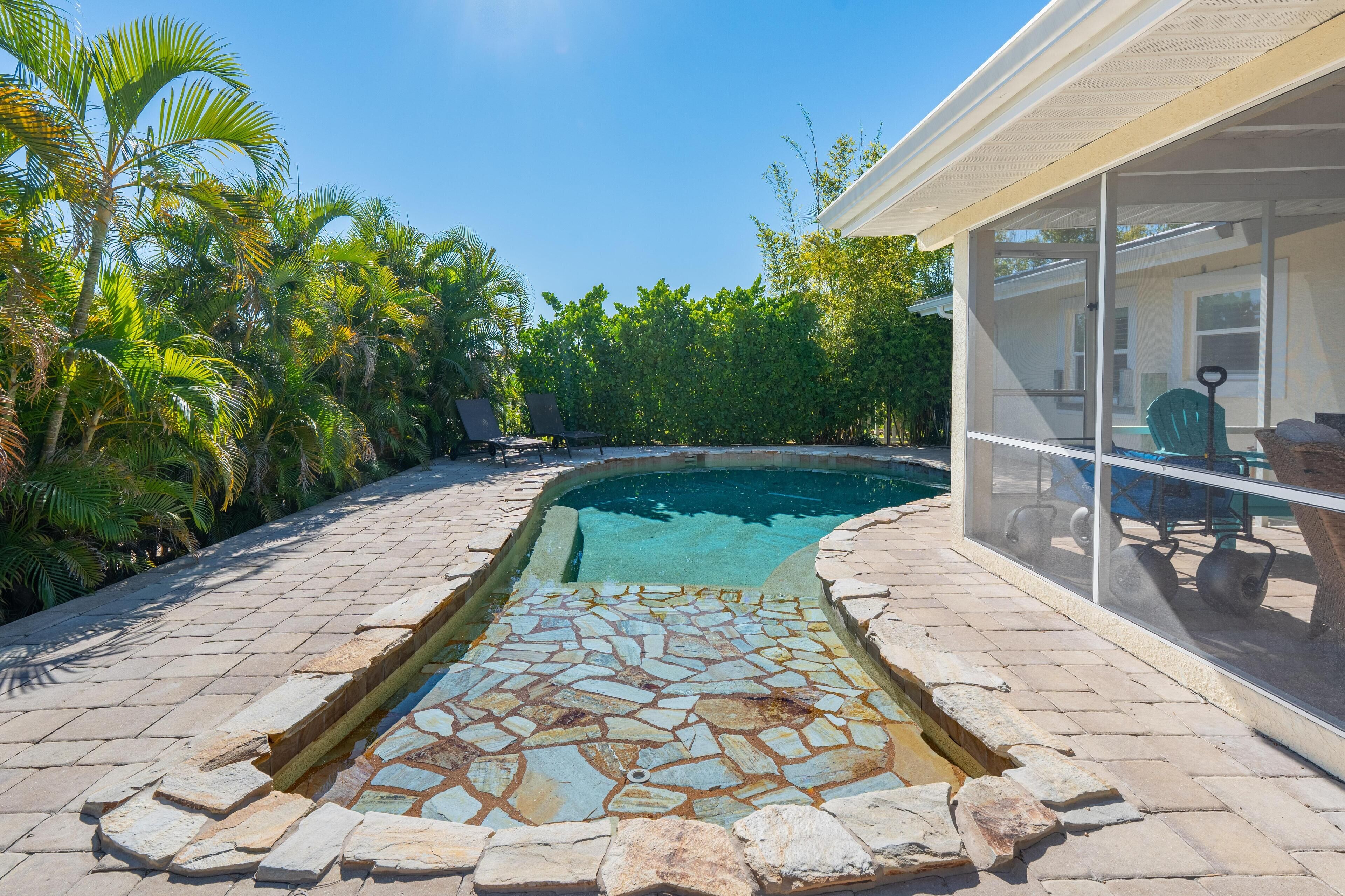Une piscine bordée d’une mosaïque de pierre, entourée de palmiers et dotée d’un patio couvert.
