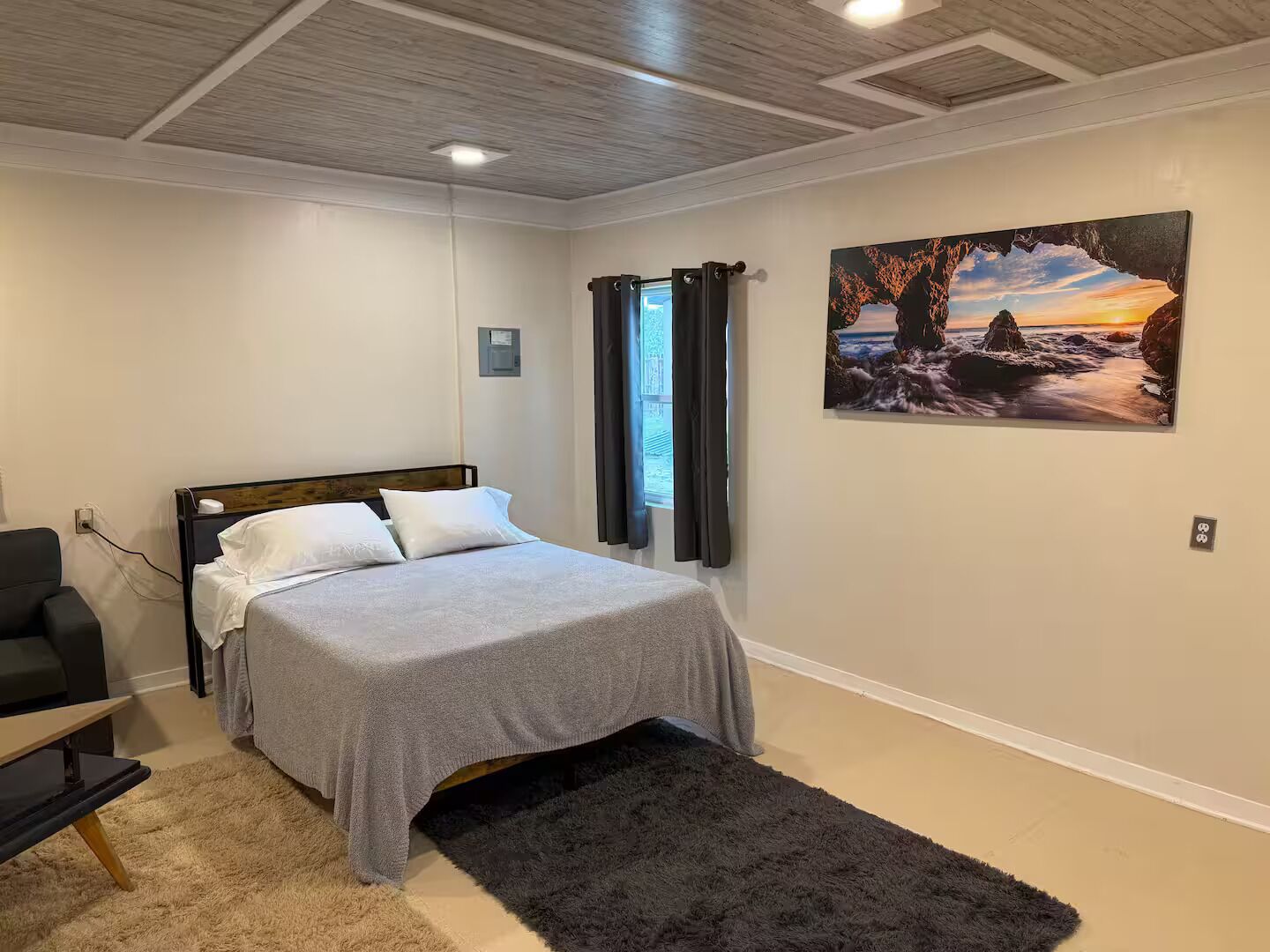 A bedroom with a bed, a desk, a chair, a painting, and a window with curtains.
