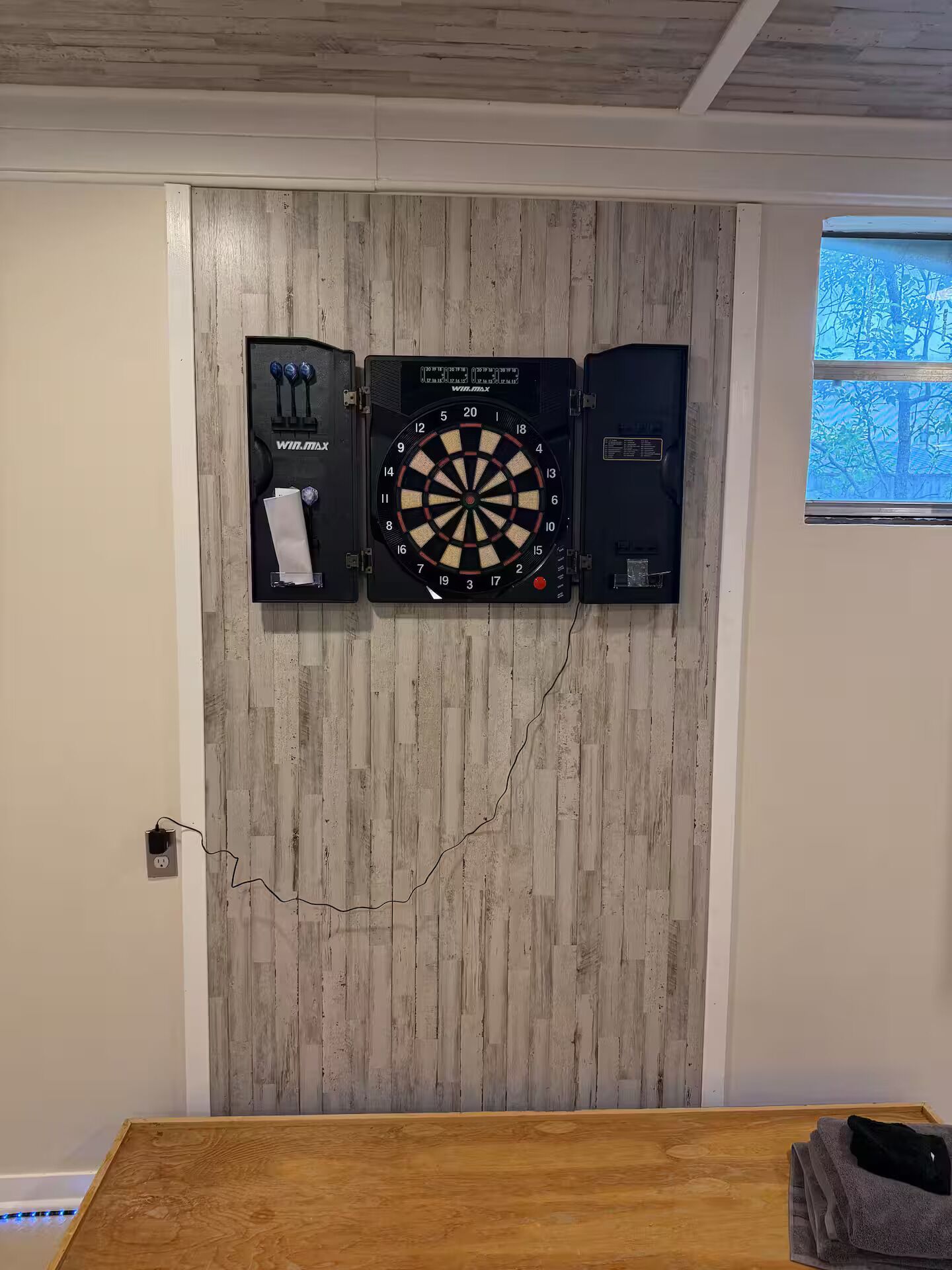 A dartboard mounted on a wooden wall with a window showing trees.