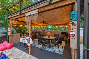 An outdoor patio area with a wooden deck, a round table with chairs, tiki bar signage, and decorative masks. - Amazing Heated & Screen Pool w/ Stunning Design! (Crystal Beach)