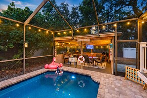 An outdoor pool area with a covered seating area, a television, and a flamingo float. - Amazing Heated & Screen Pool w/ Stunning Design! (Crystal Beach)
