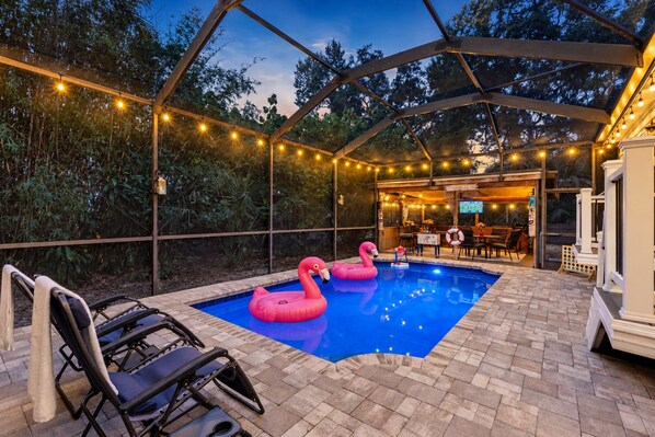 An outdoor swimming pool with two pink flamingo floats, surrounded by a covered patio with string lights, and a seating area with lounge chairs. - Amazing Heated & Screen Pool w/ Stunning Design! (Crystal Beach)