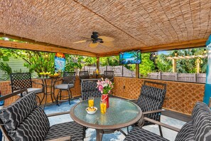 An outdoor patio with round glass tables, black and white patterned chairs, a ceiling fan, and a TV screen displaying a beach scene. - Amazing Heated & Screen Pool w/ Stunning Design! (Crystal Beach)