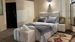 Une chambre à coucher comprenant un lit, des tables de chevet, une commode, une lampe fixée au mur, une porte et un tapis.