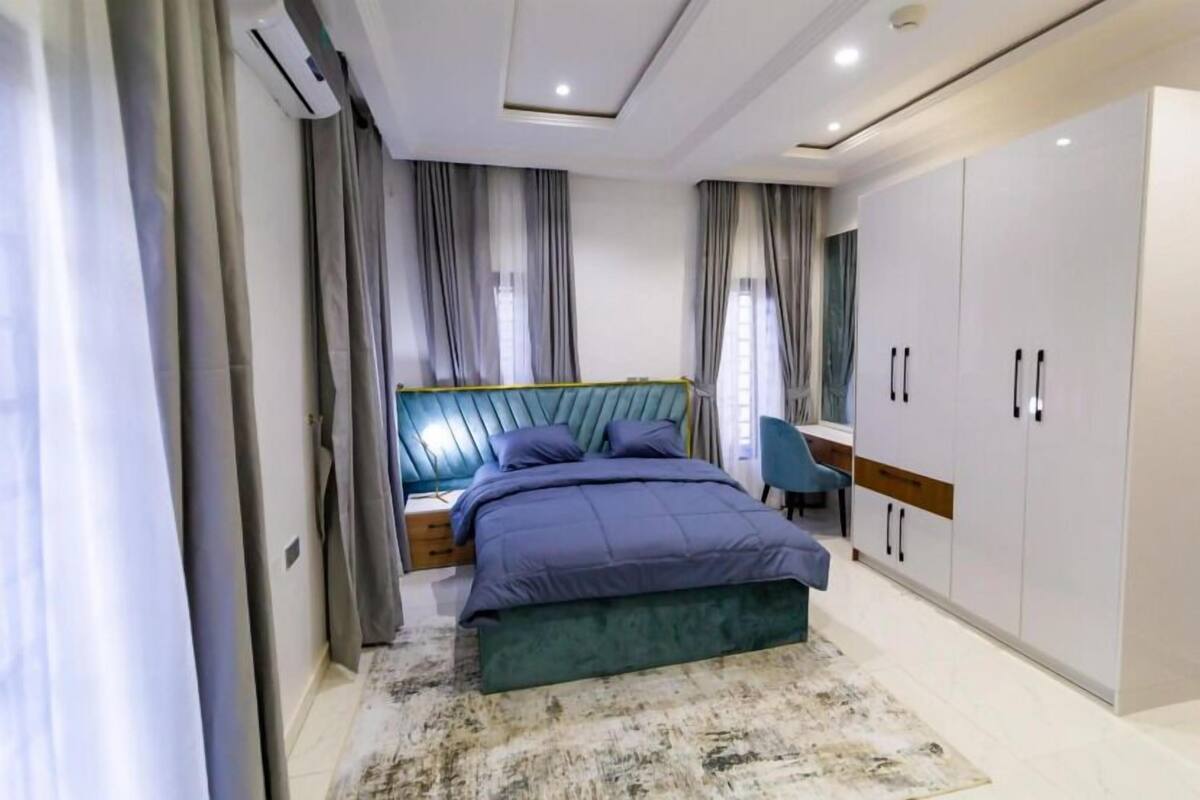 A modern bedroom with a large bed, grey curtains, a wardrobe, and a bedside table.