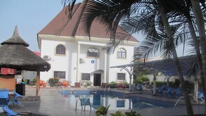 A pool area with lounge chairs, a thatched-roof structure, and a large building with a red roof. - Pauliham Hotel and Suites (Abuja)