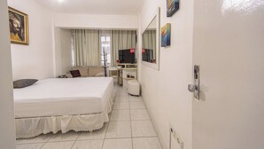 A hotel room with a bed, a desk with a computer, a TV, and a window with curtains. - Apto com Wi Fi e Garagem a 240 Metros da Praia (Santos)