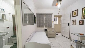 A compact hotel room with a single bed, a small sitting area, a bathroom with a sink and mirror, and a ceiling fan. - Apto com Wi Fi e Garagem a 240 Metros da Praia (Santos)