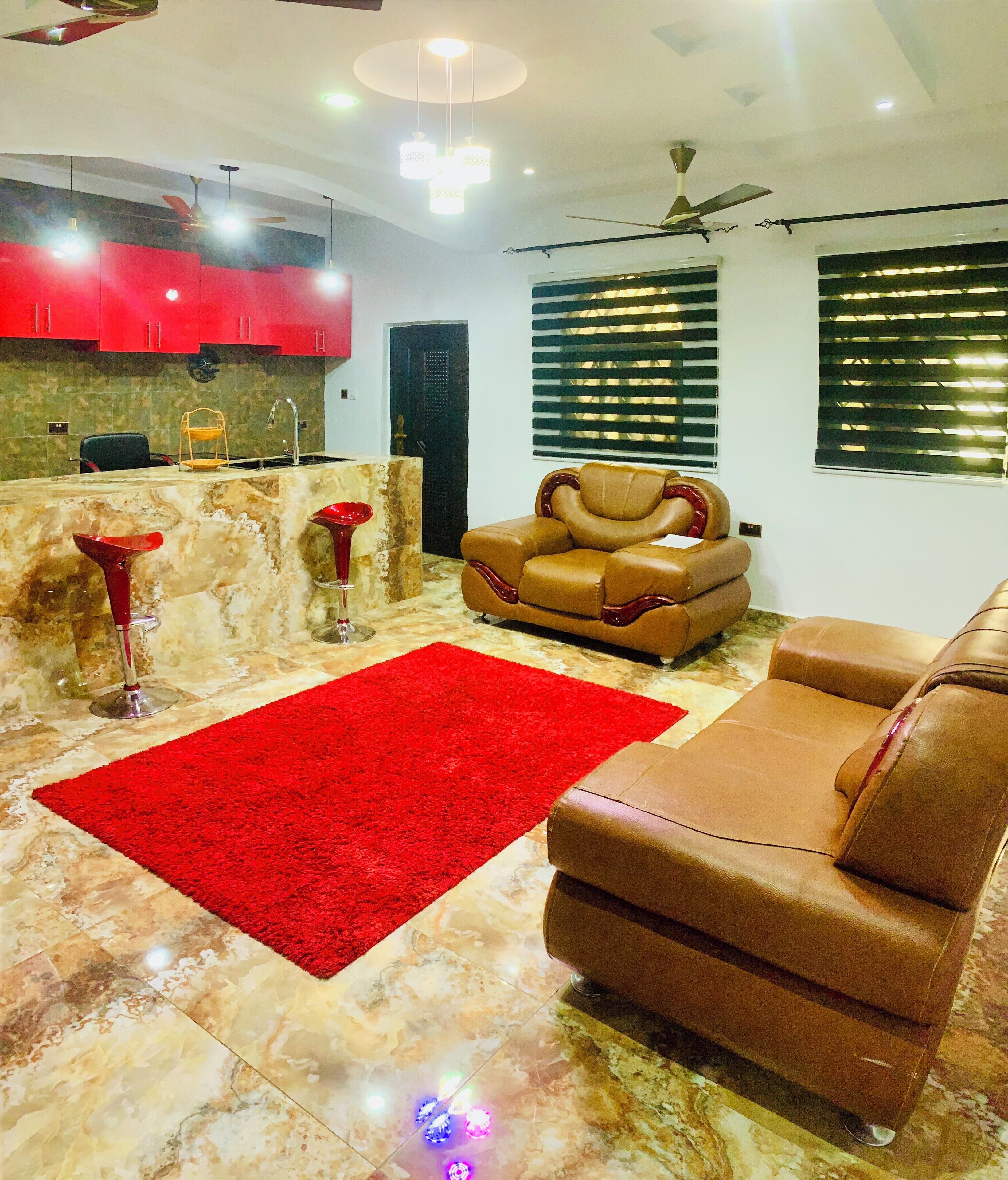 Uma sala de estar com tapete vermelho, sofá de couro marrom e área de cozinha com armários vermelhos e bancada de mármore.