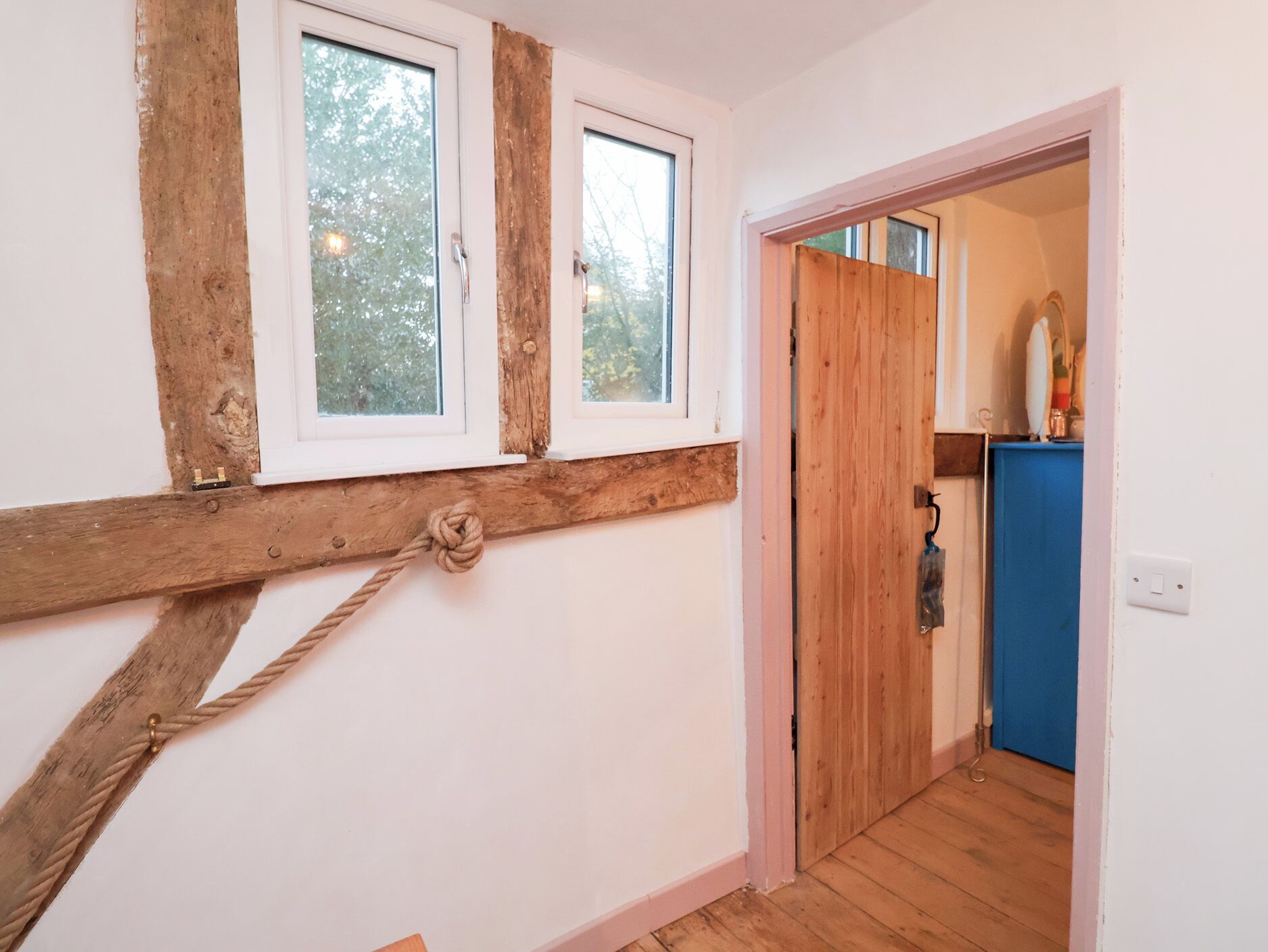 A room with wooden beams, a rope, and a door leading to a bathroom with a blue cabinet.