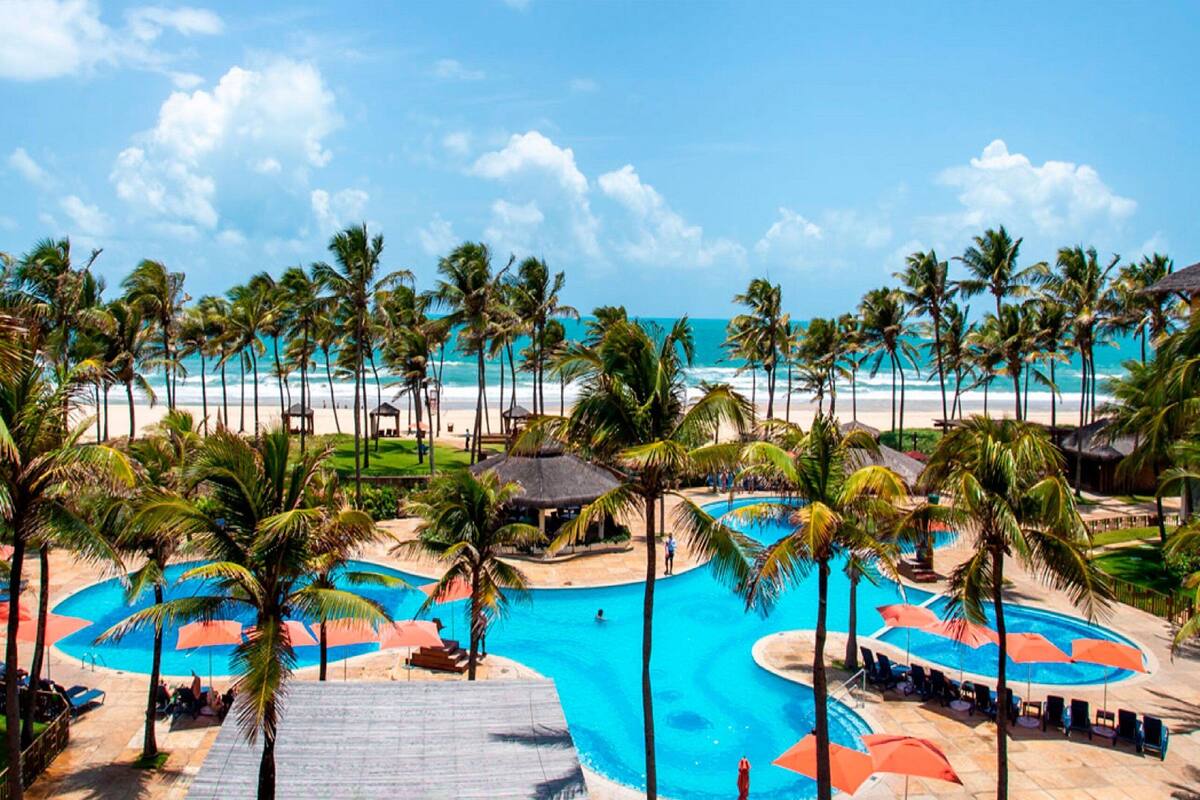 Een resort met een groot zwembad, palmbomen en uitzicht op het strand.