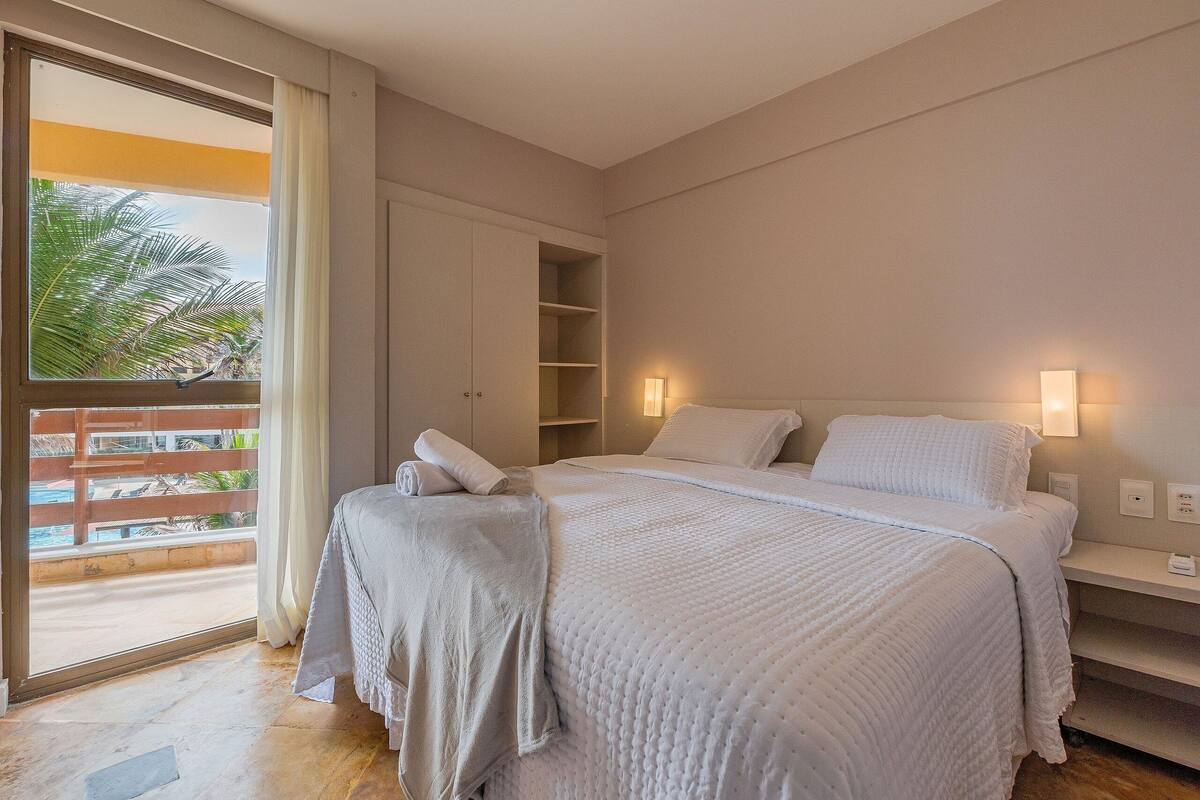 Een slaapkamer met een groot bed, een nachtkastje, een kledingkast en uitzicht op een zwembad en palmbomen door een schuifdeur.