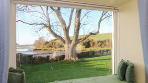 A room with a large window overlooking a grassy area and a tree. There is a bed with green bedding and pillows. - Bryncyn (Newport)