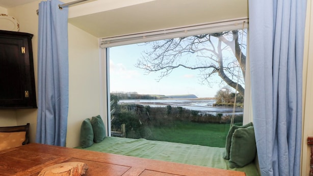 A room with a wooden table, a green cushioned bench, and a large window with a view of a landscape. - Bryncyn (Newport)