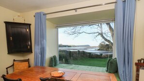 A dining room with a wooden table, chairs, and a view of a grassy area and tree through a large window. - Bryncyn (Newport)