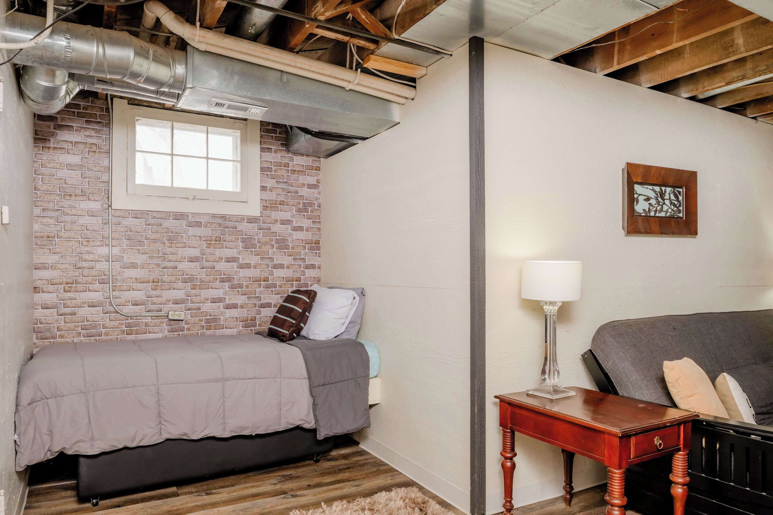 A bedroom with a brick wall, a window, a bed, a nightstand, and a sofa.