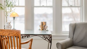 A room with a wooden chair, a glass table, a lamp, and a view of the outside through large windows. - Waterfront House w/ Patio Near Riverside Park (Kaukauna)