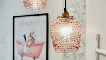 Due lampade pendenti con finiture materiche appese al soffitto, un'illustrazione incorniciata al muro e una sedia bianca con cuscino decorato.