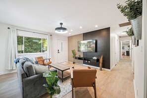 Een moderne woonkamer met een bank, salontafel en stoelen, een flatscreen televisie aan de muur en een groot raam met gordijnen.