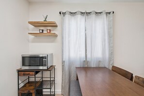 Eine kleine Küchenzeile mit einer Mikrowelle, einem Holztisch und einem Stuhl. Darüber befinden sich zwei Regale, und ein Fenster mit transparenten Vorhängen.