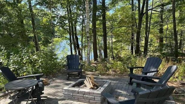 Un espacio al aire libre con sillas tipo Adirondack y una fogata, rodeado de un paisaje boscoso.