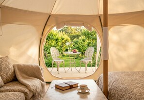 A tent with a view of a garden, two chairs, a table with a book and a cup, and a blanket on a couch.