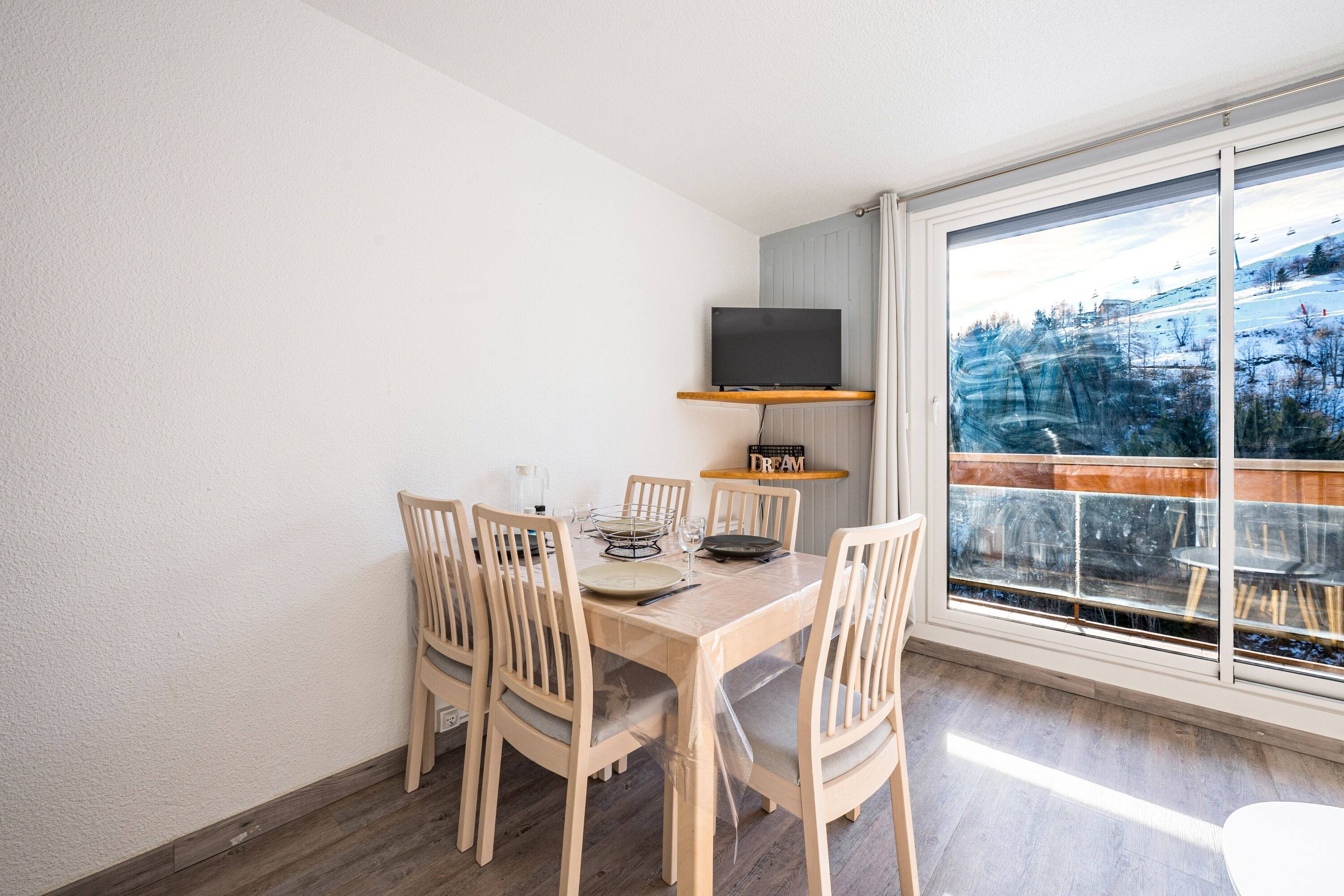 Un coin repas avec une table et des chaises en bois, un téléviseur à écran plat fixé au mur et une grande fenêtre donnant sur un paysage enneigé.