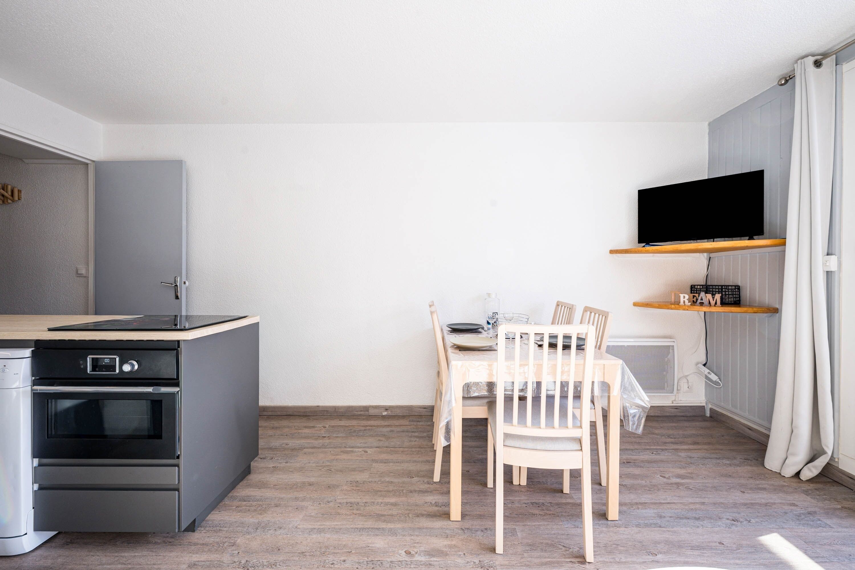 Une cuisine compacte dotée d’un four encastré, d’une table à manger avec des chaises et d’un téléviseur fixé au mur.