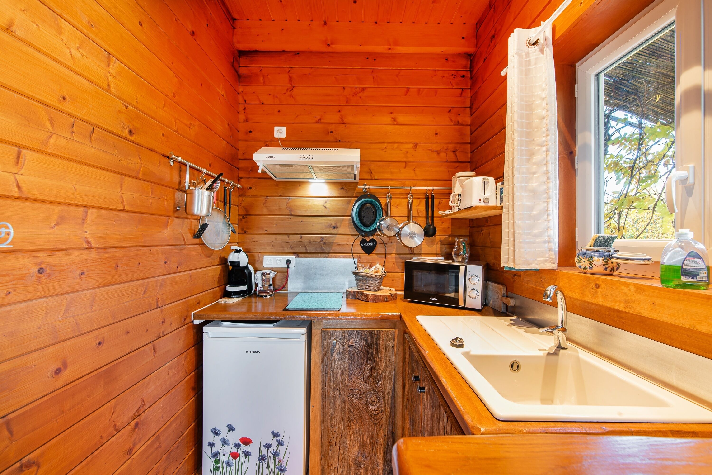 A compact kitchen with wooden walls, a refrigerator, a microwave, a sink, and a window with a view of trees.