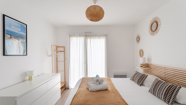Ein modernes Hotelzimmer mit einem Bett, einer weißen Kommode, einem gerahmten Bild von Saint-Malo und einem großen Fenster mit Vorhängen.