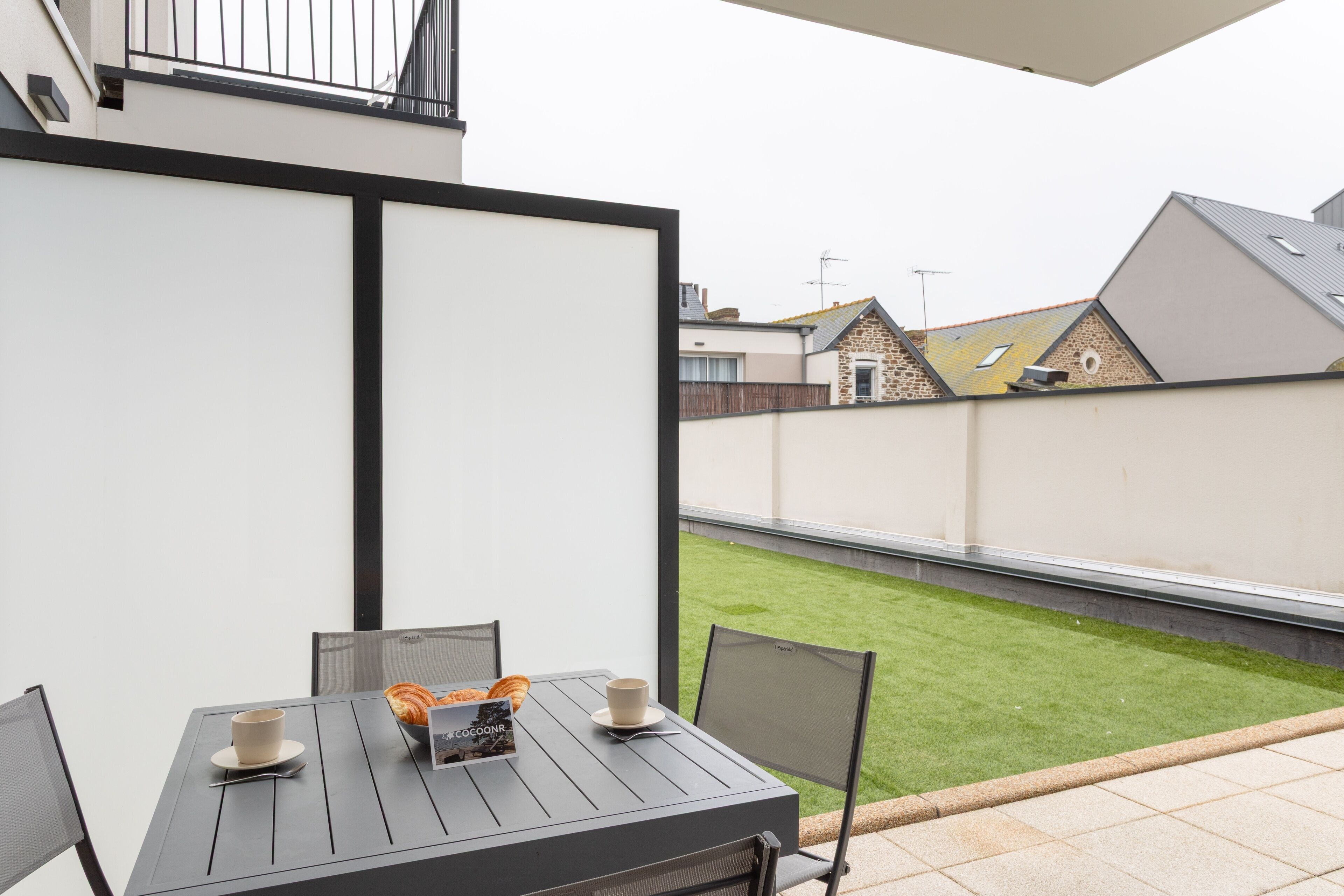 A patio with a table set for two, a chair, and a small garden visible through a fence.