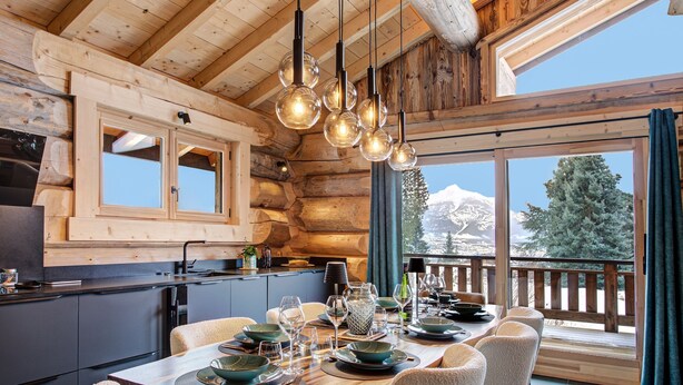 L  ’intérieur d’un chalet en bois comprend un coin repas, de grandes fenêtres et offre une vue sur une montagne enneigée.