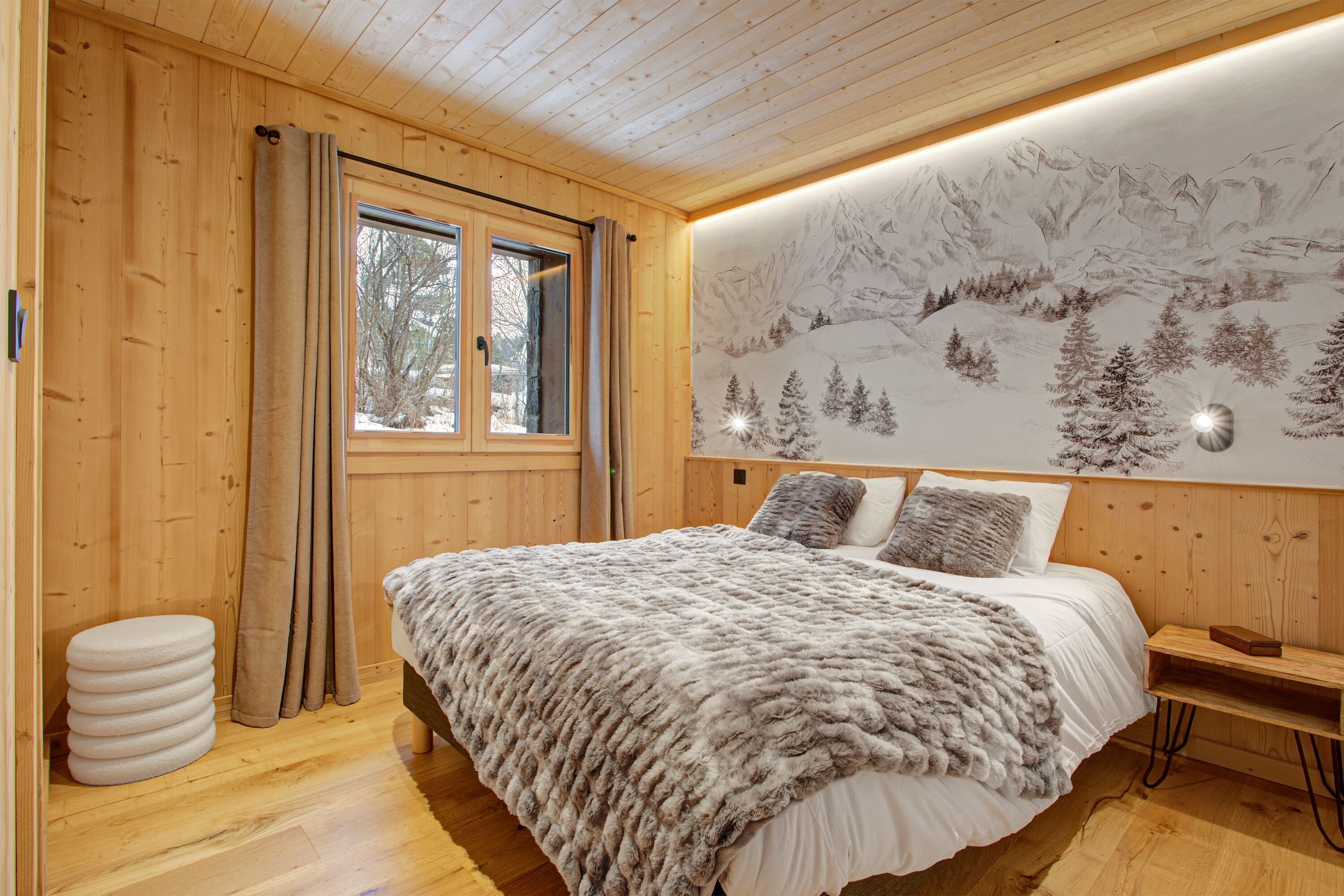 Ein Schlafzimmer einer Holzhütte mit einem Bett, einem Fenster mit Vorhängen, einer Wandmalerei einer verschneiten Berglandschaft und einem Nachttisch.