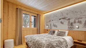 Une chambre dans une cabane en bois, avec un lit, une fenêtre avec des rideaux, une fresque murale représentant une montagne enneigée et une table de chevet.