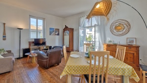 A cozy living room with a wooden floor, a dining table with chairs, a sofa, and a television. - La Villa Louise - Centre de Royan, proche plages (Royan)