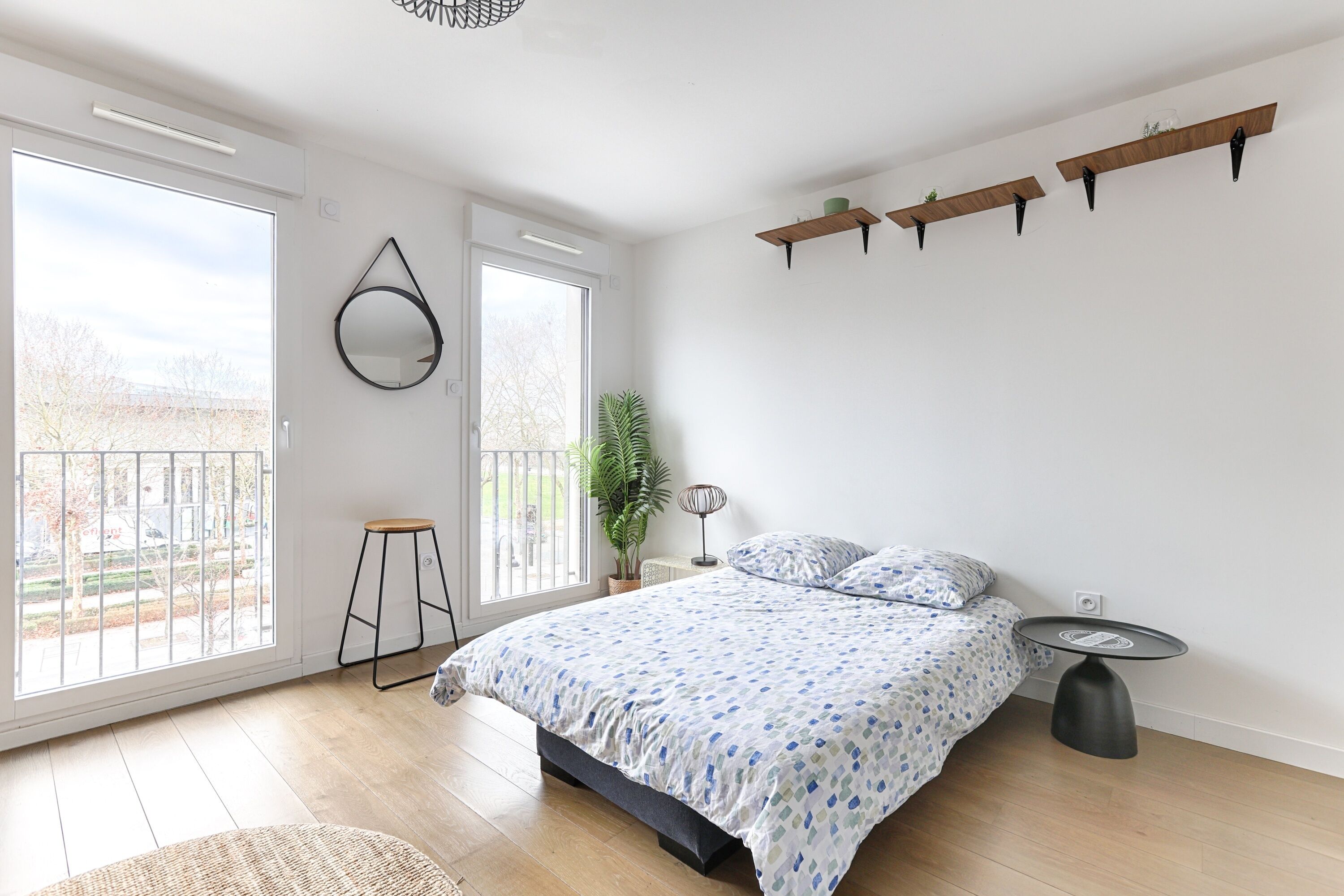 A bedroom with a large bed, a small stool, a round mirror, and a bedside table.