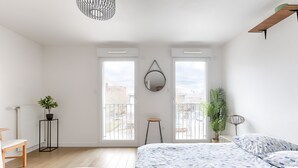 Ein modernes Schlafzimmer mit einem großen Bett, einem kleinen Hocker, einer Topfpflanze und Blick auf die Stadt durch große Fenster.