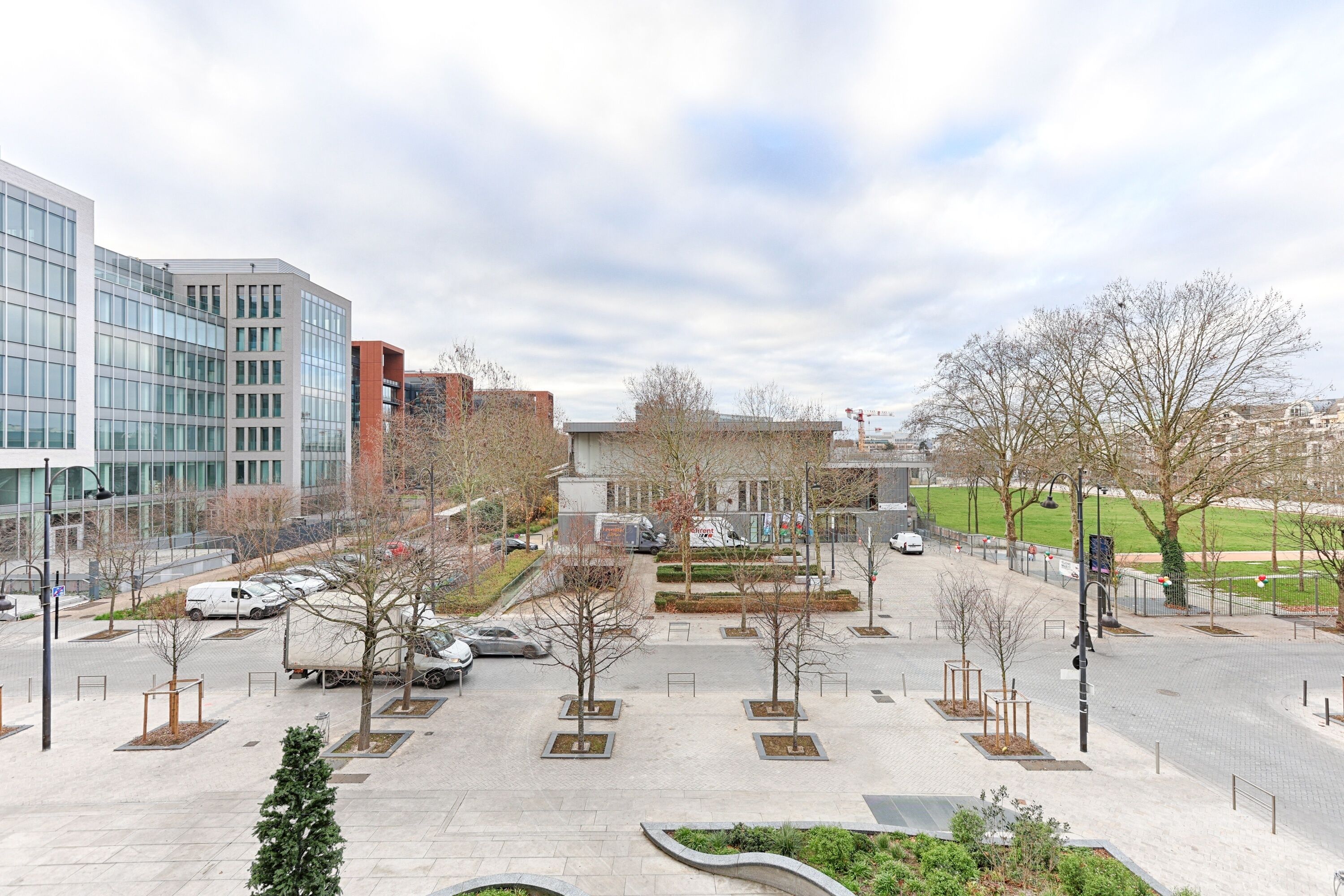 A modern urban square with a mix of commercial and residential buildings, paved walkways, and a few trees.