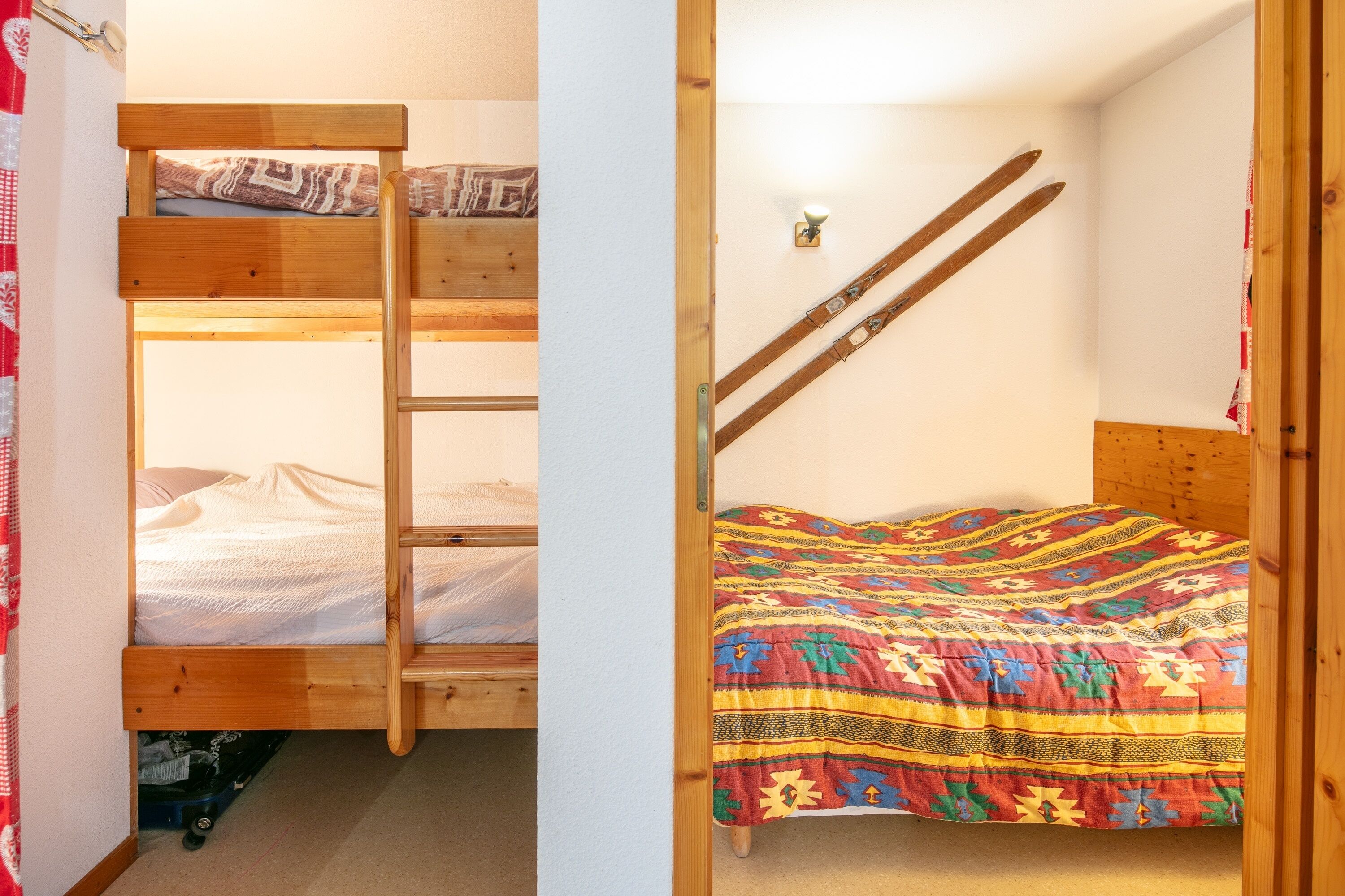 A room with a bunk bed, a lower bunk with a ladder, a colorful quilt, and a pair of skis leaning against the wall.