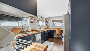Une cuisine moderne avec un plan de travail en bois, des armoires noires et une cuisinière intégrée. On y trouve deux fenêtres équipées de stores, et un réfrigérateur est visible sur la droite.