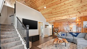 Un vaste espace de vie doté d’un plafond en bois, d’un canapé gris, d’une table basse et d’un escalier avec une rampe noire.