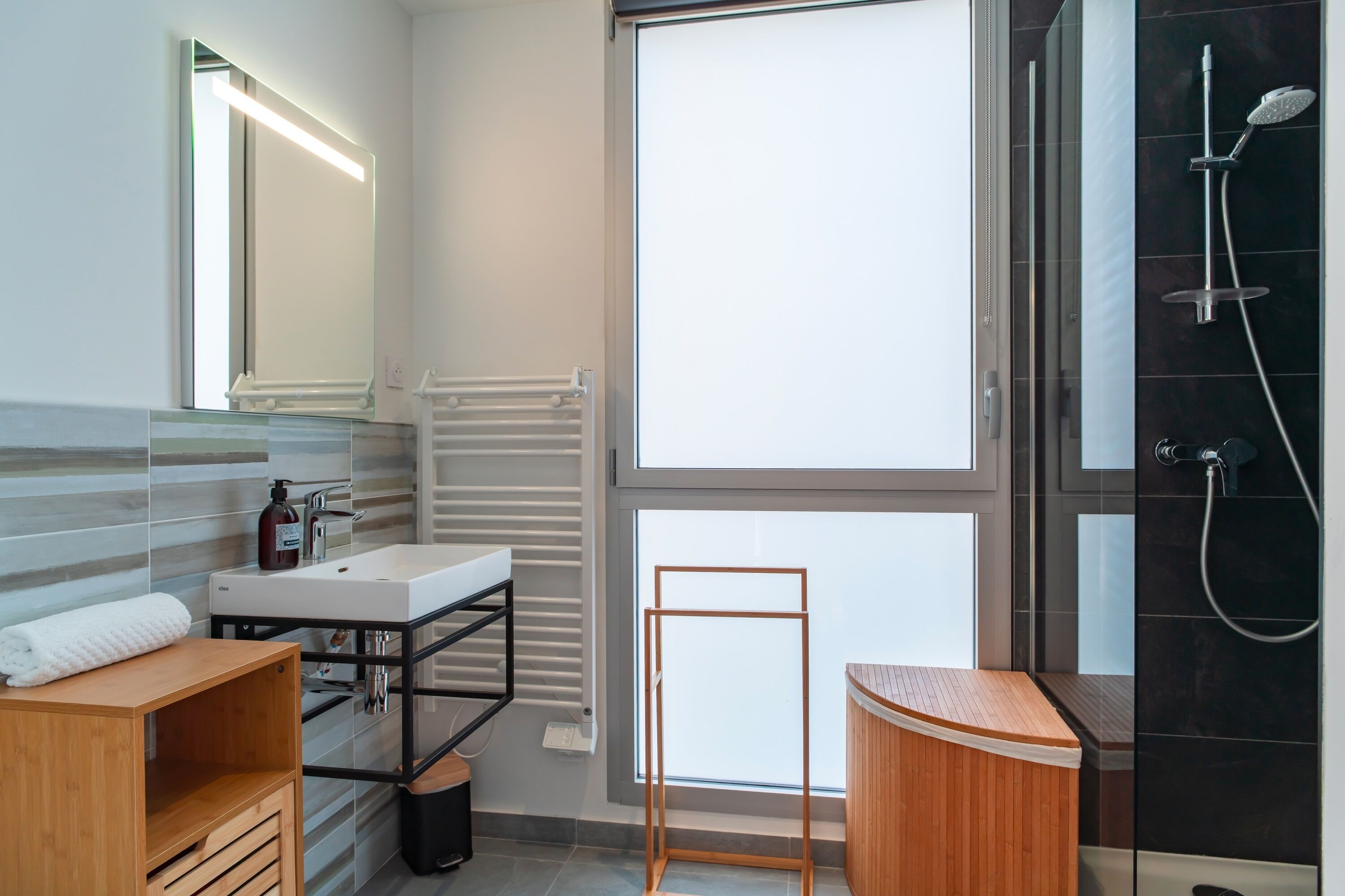 A modern bathroom with a large mirror, a white sink, a wooden vanity, a glass shower enclosure, and a window providing natural light.